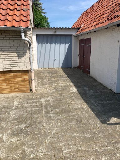 Einfahrt mit Garage und zwei Gebäuden, umgeben von Betonpflaster.